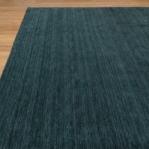 May include: A large, rectangular teal rug with a textured surface. The rug is made of a soft material and has a linear pattern. The rug is placed on a light brown wooden floor. The rug is a home decor item.