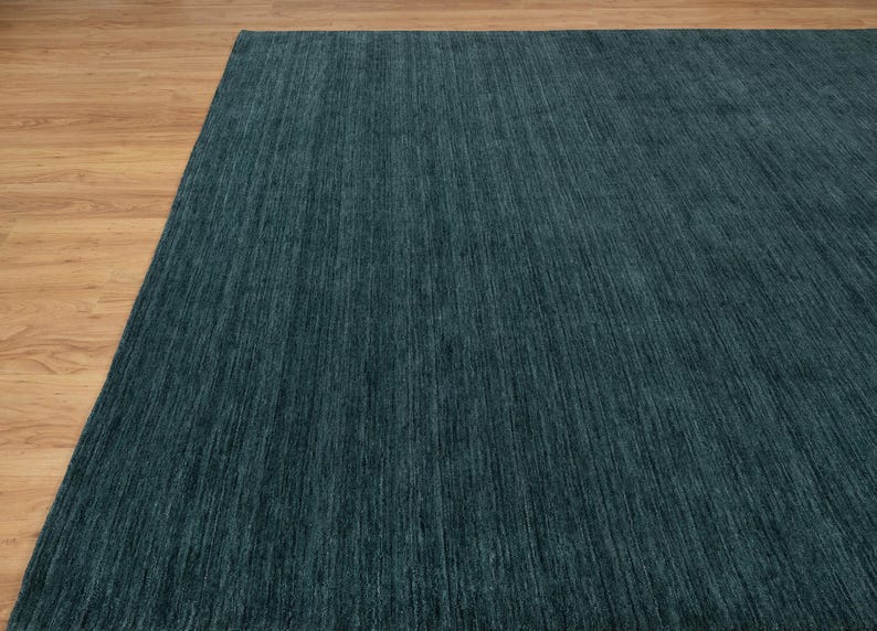 May include: A large, rectangular teal rug with a textured surface. The rug is made of a soft material and has a linear pattern. The rug is placed on a light brown wooden floor. The rug is a home decor item.