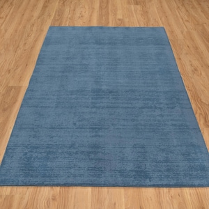 May include: A rectangular, solid blue rug with a textured surface. The rug is placed on a light brown wooden floor. The rug's color is a muted, medium blue.