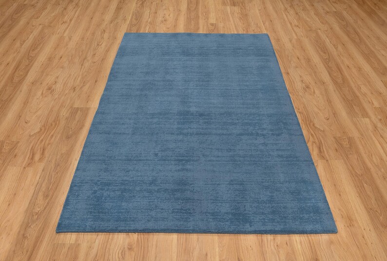 May include: A rectangular, solid blue rug with a textured surface. The rug is placed on a light brown wooden floor. The rug's color is a muted, medium blue.