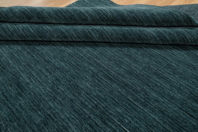 May include: A folded section of teal-colored fabric with a textured, diagonal pattern. The fabric appears to be thick and soft, with visible folds and shadows. The material is a solid teal color, with a subtle sheen.