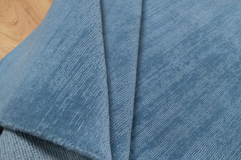 May include: Close-up of a folded, textured fabric in a muted blue hue. The material has a subtle diagonal pattern, creating a sense of depth and visual interest. The fabric's texture and color suggest it could be used for upholstery or home decor.
