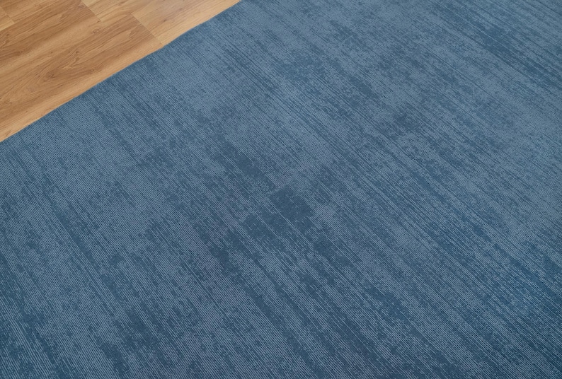 May include: A section of a blue rug with a textured surface. The rug has a diagonal pattern and is adjacent to a section of light brown wooden flooring.