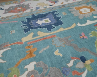 May include: A large area rug featuring a vibrant turquoise blue background with an abstract pattern of scattered geometric shapes in shades of navy blue, orange, yellow, green, pink, and gray, arranged in a non-repeating, multi-colored design.