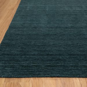May include: A section of a teal-colored rug with a textured surface. The rug is placed on a light brown wooden floor. The rug's color is a deep, saturated teal, and the texture appears to be a low pile.