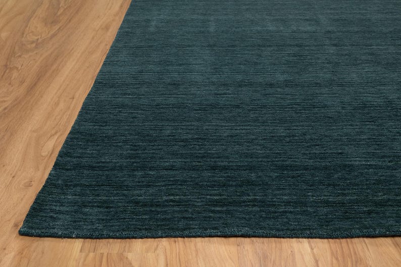 May include: A section of a teal-colored rug with a textured surface. The rug is placed on a light brown wooden floor. The rug's color is a deep, saturated teal, and the texture appears to be a low pile.