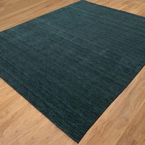 May include: A rectangular, teal-colored area rug with a textured surface. The rug is placed on a light brown wooden floor, creating a contrast in color and texture. The rug's fibers appear to run in a linear pattern.