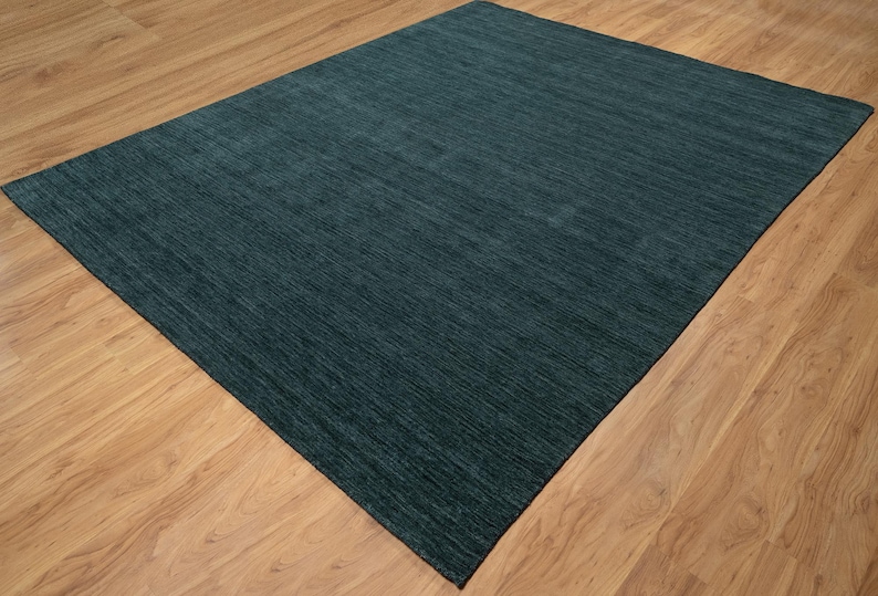 May include: A rectangular, teal-colored area rug with a textured surface. The rug is placed on a light brown wooden floor, creating a contrast in color and texture. The rug's fibers appear to run in a linear pattern.
