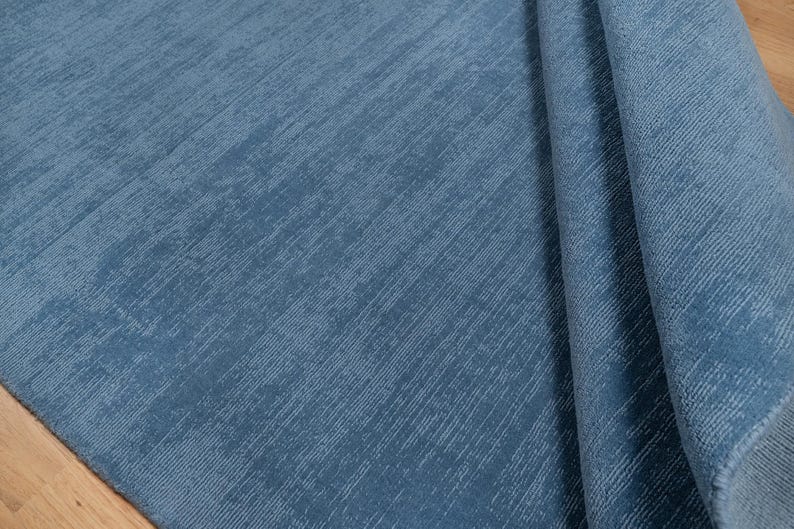 May include: A close-up of a blue rug with a textured surface. The rug has a soft, plush appearance and is folded over, revealing the underside. The color is a muted, medium blue.