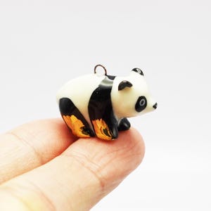 May include: A small, three-dimensional panda charm featuring a glossy white and black finish with metallic gold accents on the lower body and legs. The panda is depicted in a standing position and includes a small metal loop at the top for hanging.