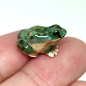 May include: A small ceramic frog figurine. The frog is primarily green with brown spots on its back and a cream-colored belly. It has black eyes and is posed in a sitting position. The figurine is held between two fingers.