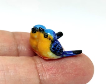 May include: A miniature ceramic sculpture of two bluebirds perched together. The birds have blue heads, wings, and tails, with yellow chests and bellies. The sculpture is detailed with black eyes and beaks.