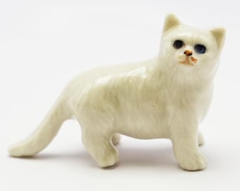 Puede incluir: Una pequeña figura de gato de cerámica blanca con textura de pelo largo, orejas puntiagudas y heterocromía, con un ojo marrón oscuro y otro azul brillante, y una pequeña mancha naranja-marrón en la zona de la nariz y la boca.