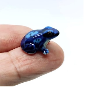 May include: A small, ceramic frog figurine in shades of blue. The frog has a glossy finish and is detailed with darker blue spots. The figurine is resting on a finger, showcasing its size and detail. The frog is a decorative item.