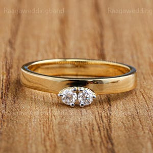 Puede incluir: Una alianza de boda dorada con dos diamantes redondos y claros, colocados uno al lado del otro. El anillo tiene una banda plana y pulida. El texto "Raagaaweddingband" es visible en la imagen. Se encuentra sobre madera.