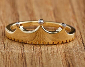 May include: A gold-colored crown ring with a detailed design. The ring features a crown shape with small silver accents on top. The ring is set against a wooden background.