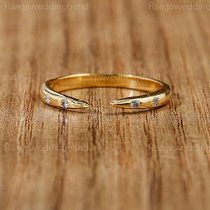 Può includere: Un anello color oro con un design aperto, con tre piccole pietre preziose trasparenti. L'anello ha una superficie liscia e lucida ed è presentato su uno sfondo di legno caldo. Il design dell'anello è moderno ed elegante.