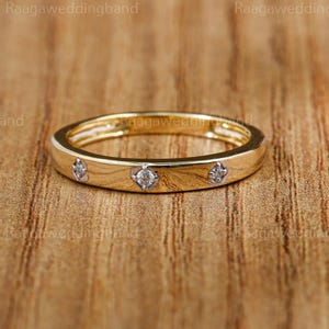 Può includere: Un anello a fascia in oro con tre piccole gemme trasparenti incastonate nella fascia. L'anello è su una superficie di legno. Il testo "Raaga wedding band" è visibile sullo sfondo.