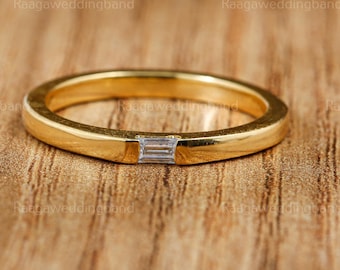 May include: A gold-colored ring with a rectangular, clear gemstone set in the center. The ring is on a wooden surface. The text "RaagaWeddingband" is repeated around the ring.