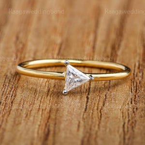 Peut inclure: Une bague dorée ornée d'une pierre précieuse triangulaire et transparente. La bague présente un anneau fin et un design simple, sur un fond en bois chaleureux. La pierre précieuse est le point central, captant la lumière.