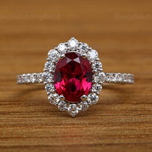 May include: A silver ring featuring a large, oval-cut, deep red gemstone, encircled by a halo of smaller, clear gemstones. The ring's band is also set with small, clear gemstones. The ring is displayed against a warm, wooden backdrop.