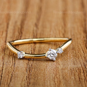 May include: A curved gold ring with three clear gemstones. The ring has a delicate design and is set against a wooden background. The text "RaagaWeddingband" is visible in the background.