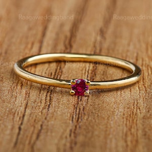 Puede incluir: Un anillo de oro con una pequeña gema roja cuadrada en el centro. El anillo es delgado y delicado, con un acabado pulido. La gema es de un color rojo vibrante, y el anillo está sobre un fondo de madera.