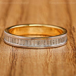 May include: A gold and silver wedding band with a row of rectangular-cut stones. The ring has a gold interior and a silver exterior with the stones set in a channel. The band is on a wooden surface.
