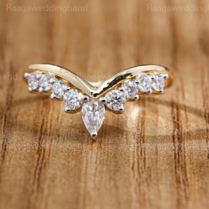 Puede incluir: Un anillo dorado en forma de V con múltiples piedras redondas y transparentes, y una piedra en forma de pera en el centro. El anillo se encuentra sobre un fondo de madera, resaltando su diseño y brillo.