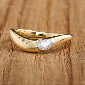 May include: A gold ring featuring a marquise-cut diamond set in a white metal bezel. The ring has a curved, contemporary design. The background is a wooden surface.