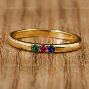 Puede incluir: Un anillo de banda de oro con tres piedras preciosas: verde esmeralda, magenta y azul zafiro. El anillo está sobre una superficie de madera. El texto "Raaga wedding band" es visible en el fondo.