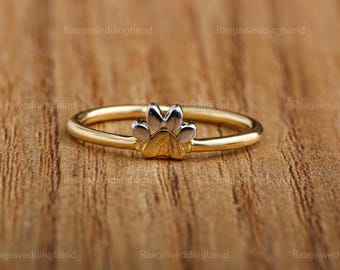Puede incluir: Un anillo dorado con un diseño de huella de pata. La huella de pata es bicolor, con una base dorada y detalles plateados. El anillo está sobre un fondo de madera.