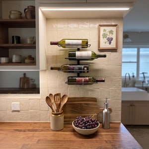 Pode incluir: Uma cena de cozinha com uma bancada de madeira, apresentando um suporte para vinho com quatro garrafas. Uma tigela de uvas, uma tábua de cortar e utensílios em um pote de cerâmica também estão presentes. Uma impressão botânica emoldurada e um dispensador de óleo de aço inoxidável completam a configuração.
