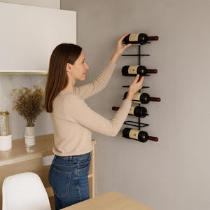May include: A black metal wall-mounted wine rack holding several bottles of red wine. The rack is mounted on a gray wall. A person is placing a bottle into the rack. The bottles have dark glass and red wine.
