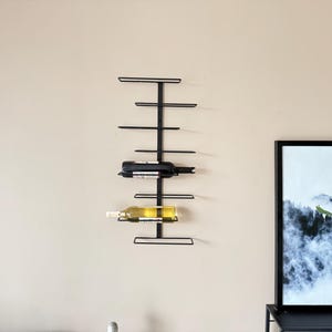 May include: A black metal wall-mounted wine rack holding two wine bottles. The rack has a vertical design with horizontal bars to hold the bottles. The bottles are a dark red and a light yellow color. The rack is mounted on a beige wall.