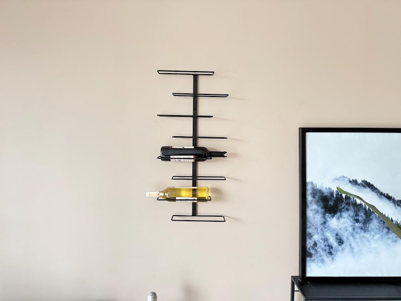 May include: A black metal wall-mounted wine rack holding two wine bottles. The rack has a vertical design with horizontal bars to hold the bottles. The bottles are a dark red and a light yellow color. The rack is mounted on a beige wall.
