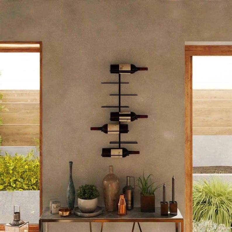 May include: A black metal wall-mounted wine rack holding four wine bottles. The rack is mounted on a gray wall. The bottles are displayed horizontally. A table with decorative items is below the rack.