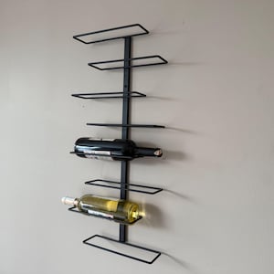 May include: A black metal wall-mounted wine rack with multiple horizontal shelves. Two wine bottles are displayed, one red and one white. The rack is mounted on a neutral-colored wall, providing a space-saving storage solution for wine bottles.