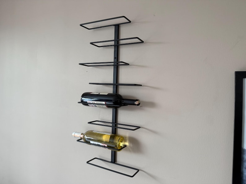 May include: A black metal wall-mounted wine rack with multiple horizontal shelves. Two wine bottles are displayed, one red and one white. The rack is mounted on a neutral-colored wall, providing a space-saving storage solution for wine bottles.