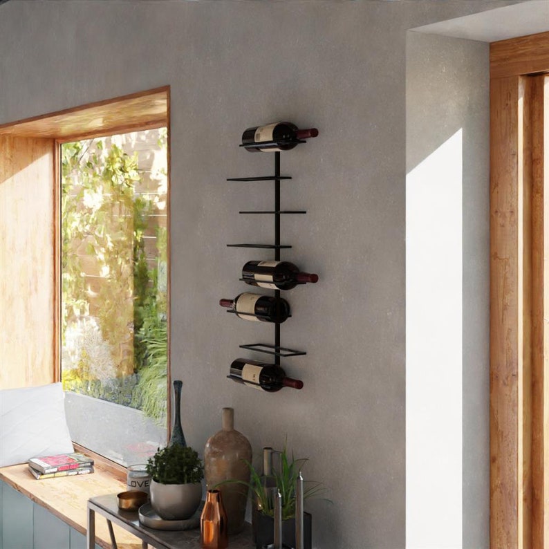 May include: A black metal wall-mounted wine rack holding several bottles of red wine. The rack has a vertical design with horizontal shelves to hold the bottles. The wine rack is mounted on a gray wall near a window.
