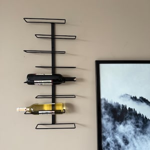 May include: A black metal wall-mounted wine rack holding two wine bottles. The rack has a vertical support with horizontal bars to hold the bottles. The bottles are a dark red and a light yellow color. The rack is mounted on a beige wall.