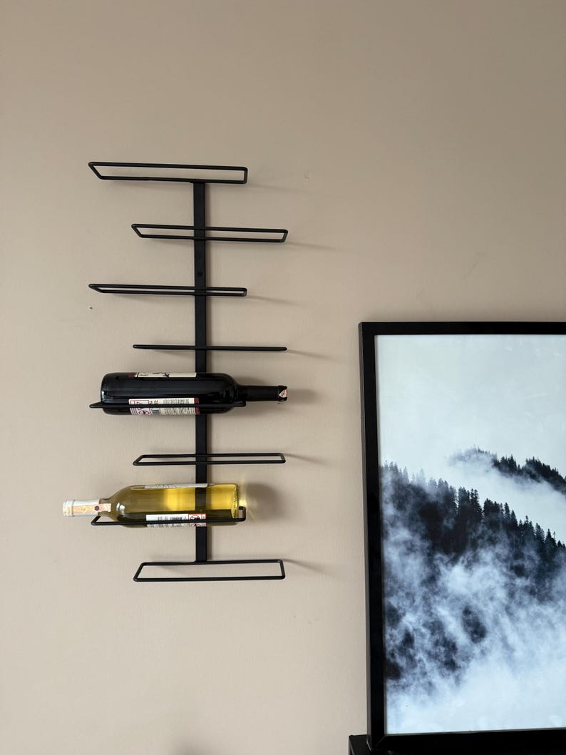 May include: A black metal wall-mounted wine rack holding two wine bottles. The rack has a vertical support with horizontal bars to hold the bottles. The bottles are a dark red and a light yellow color. The rack is mounted on a beige wall.