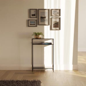 Könnte beinhalten: Ein schwarzer Konsolentisch aus Metall mit zwei Ablagen. Der Tisch steht in einem Raum mit weißen Wänden und einem Holzboden. Über dem Tisch hängen mehrere gerahmte Bilder an der Wand.
