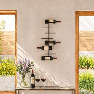 May include: A black metal wall-mounted wine rack holding four bottles of wine. The rack has a minimalist design with horizontal bars to support the bottles. The bottles are displayed against a light gray wall, with a table and wine glasses in the foreground.
