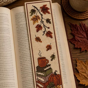 Può includere: Un segnalibro rettangolare a punto croce raffigurante un ramo verticale con foglie autunnali nei toni del rosso, arancione e verde. Sotto il ramo, una pila di libri è sormontata da una tazza fumante e da una zucca arancione, tutto a punto croce.