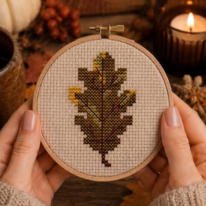 May include: A circular wooden embroidery hoop containing a piece of beige Aida fabric with a cross-stitch design of an oak leaf. The leaf features a gradient of green, yellow, and dark brown thread colours, centred within the frame.