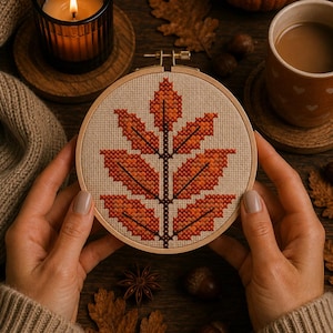 May include: A circular wooden embroidery hoop containing a cross-stitch pattern of a single large autumn leaf. The leaf is composed of various shades of orange, red, and brown thread stitched onto a light beige fabric background.