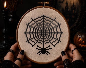 May include: A circular wooden embroidery hoop containing a piece of beige fabric with a black cross-stitch design of a spider web and a small spider hanging from the center. The design is centered on the fabric within the round frame.