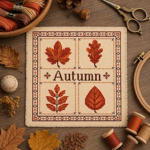 May include: A square cross-stitch sampler on cream fabric featuring four distinct autumn leaves in shades of orange, red, and brown, arranged in a grid. The word Autumn is stitched in dark brown letters in the centre, surrounded by a geometric border.