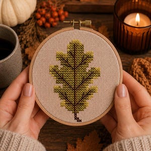 May include: A circular wooden embroidery hoop containing a cross-stitch design of a single oak leaf. The leaf is rendered in shades of green and yellow with dark brown stitching for the veins, set against a light beige fabric background.
